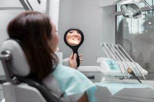 770457 - Riberdental - Sonajero.es Patient in a dental chair smiles at their reflection in a handheld mirror during a dental visit with dental tools nearby: tray and overhead light visible.