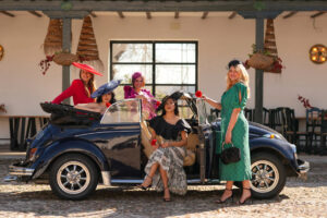 772931 - DNUE for Ladies 1 - Sonajero.es Group of women in colorful outfits and wide-brim hats posing with a dark blue vintage convertible in a sunlit cobblestone courtyard.