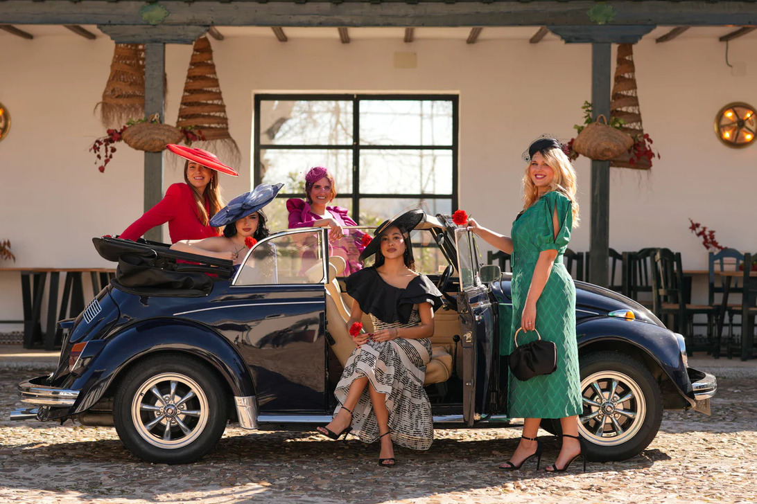 772931 - DNUE for Ladies 1 - Sonajero.es Group of women in colorful outfits and wide-brim hats posing with a dark blue vintage convertible in a sunlit cobblestone courtyard.