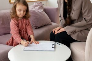 773137 - Garo Psicología - Sonajero.es Little girl drawing on a clipboard at a small round table while a caregiver sits nearby on a couch.