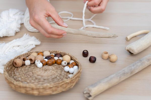 Hands string wooden beads on a cord for a bracelet or necklace, with a basket of beads nearby and skeins of cord on a wooden table.
