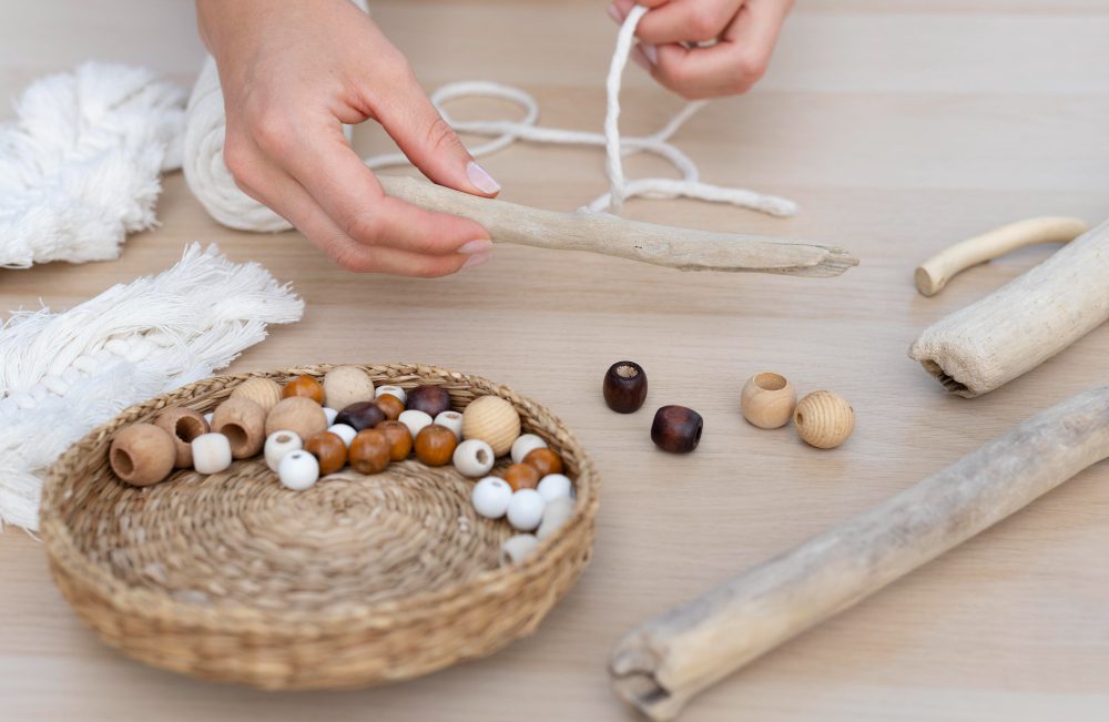 775287 - Horizonte Talleres Waldorf - Sonajero.es Hands string wooden beads on a cord for a bracelet or necklace, with a basket of beads nearby and skeins of cord on a wooden table.
