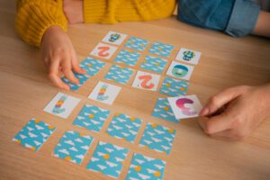 Two players sit at a wooden table playing a memory card game with blue cloud-pattern cards laid out in a grid and a few cards being flipped.