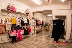 Children's clothing store interior with racks of colorful outfits, shelves, and a light wood floor.