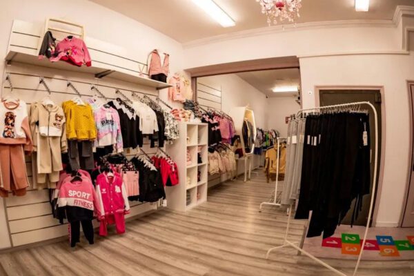 Children's clothing store interior with racks of colorful outfits, shelves, and a light wood floor.