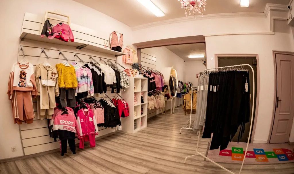 777655 - Yarelis - Sonajero.es Children's clothing store interior with racks of colorful outfits, shelves, and a light wood floor.