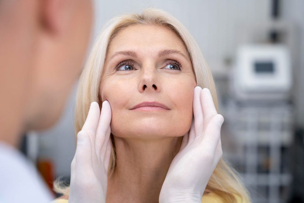 780480 - Catalano Medicina Estética - Sonajero.es Healthcare provider wearing gloves examines a middle-aged woman's face in a clinical setting.